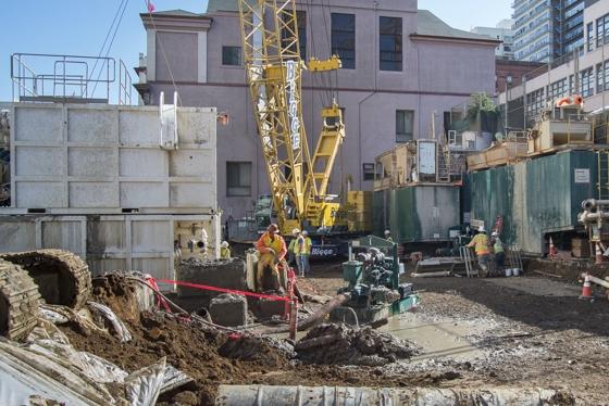 Construction equipment and crews work at the future Chinatown Station site surrounded by concrete barriers and buildings.