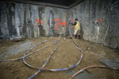 Worker in yellow vest preparing soil grouting equipment in underground transit station concourse with exposed concrete walls.