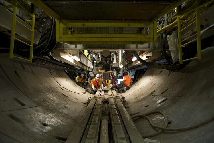 Workers in orange vests inside underground tunnel with yellow overhead crane and concrete tunnel segments.