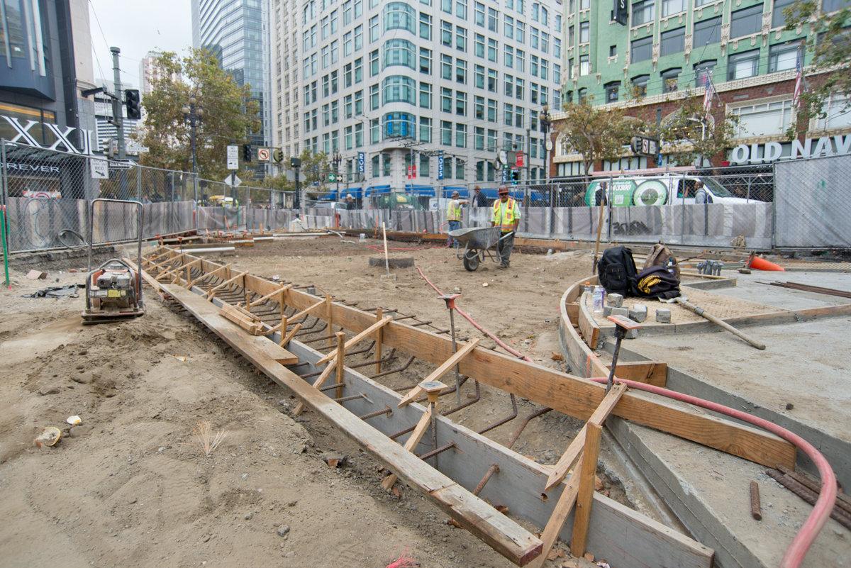 Construction site with wooden forms for concrete foundation at urban intersection surrounded by tall buildings.
