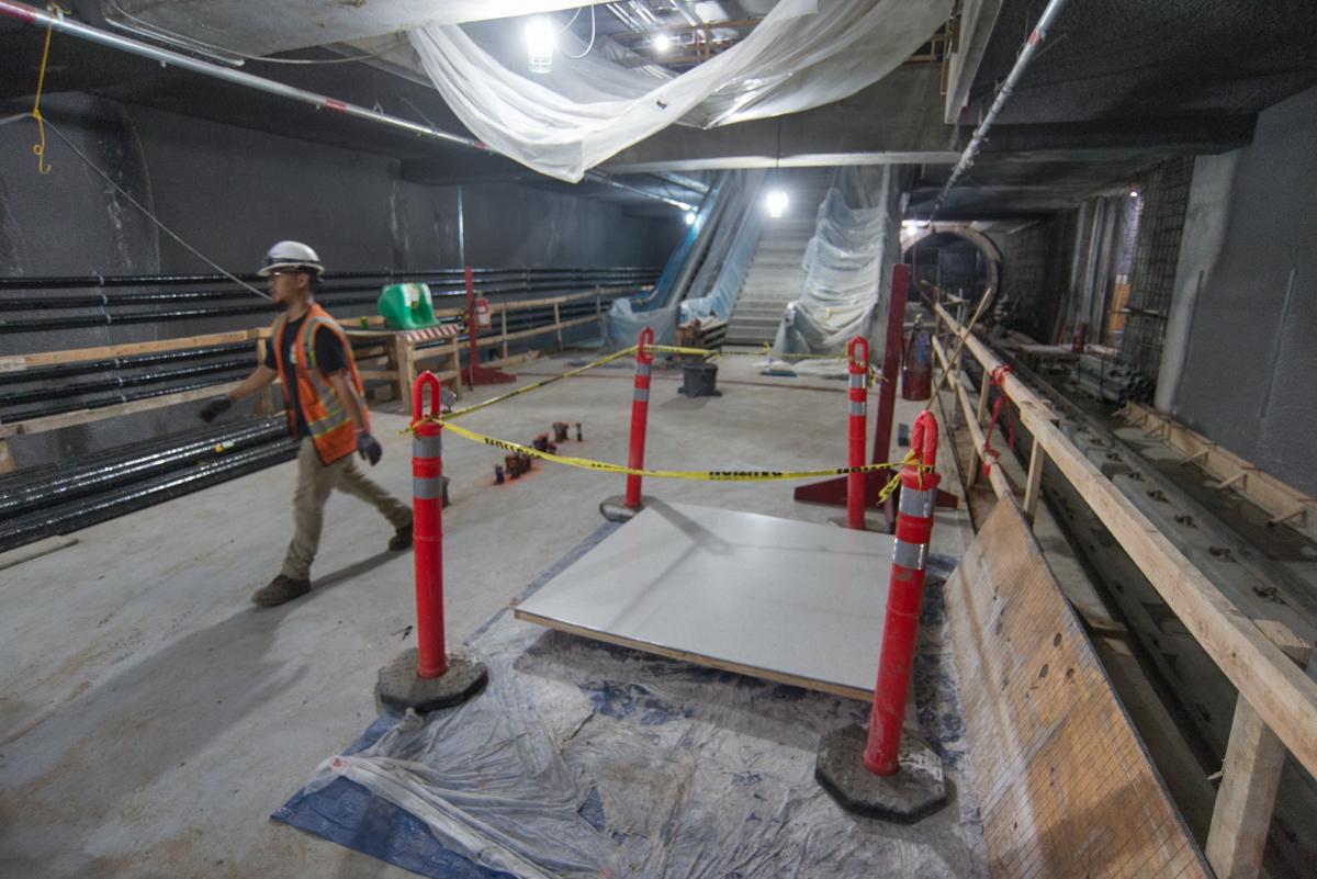 Worker in hard hat and safety vest inspects terrazzo flooring test section in underground transit station during night construction.