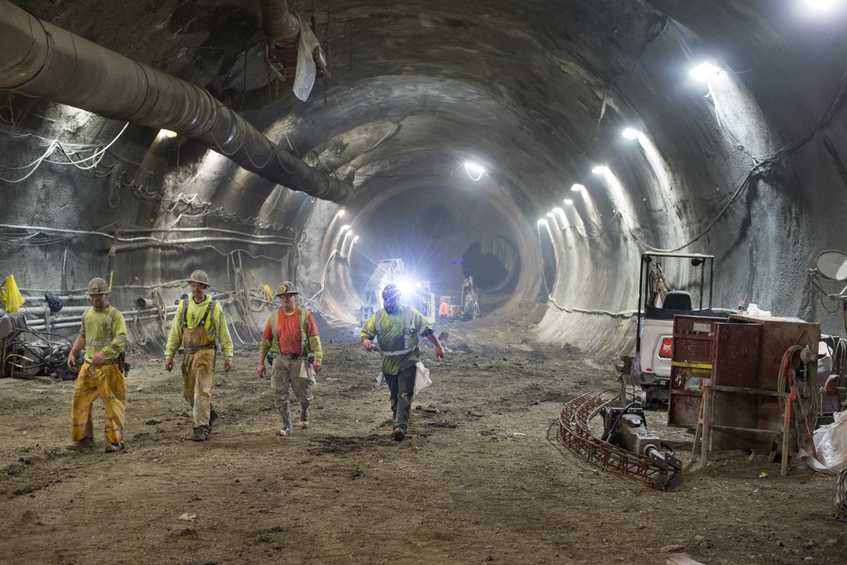 Workers in safety vests walk through an underground tunnel under construction with bright lighting and equipment visible.