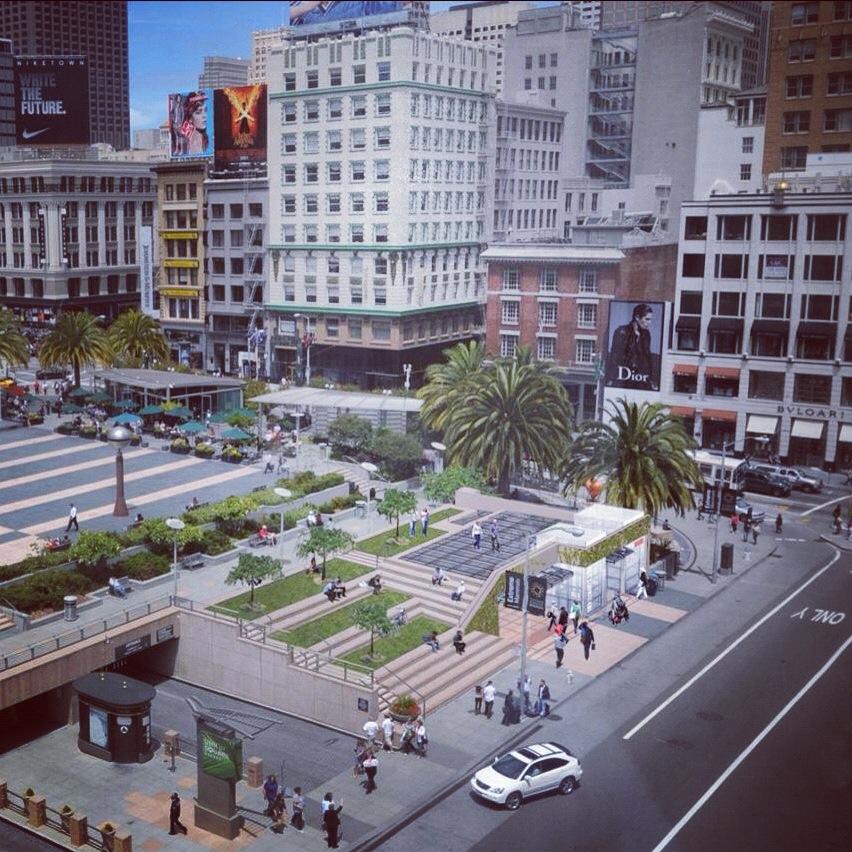 Union Square plaza with palm trees, pedestrians, surrounding buildings, and street-level retail areas in downtown San Francisco.