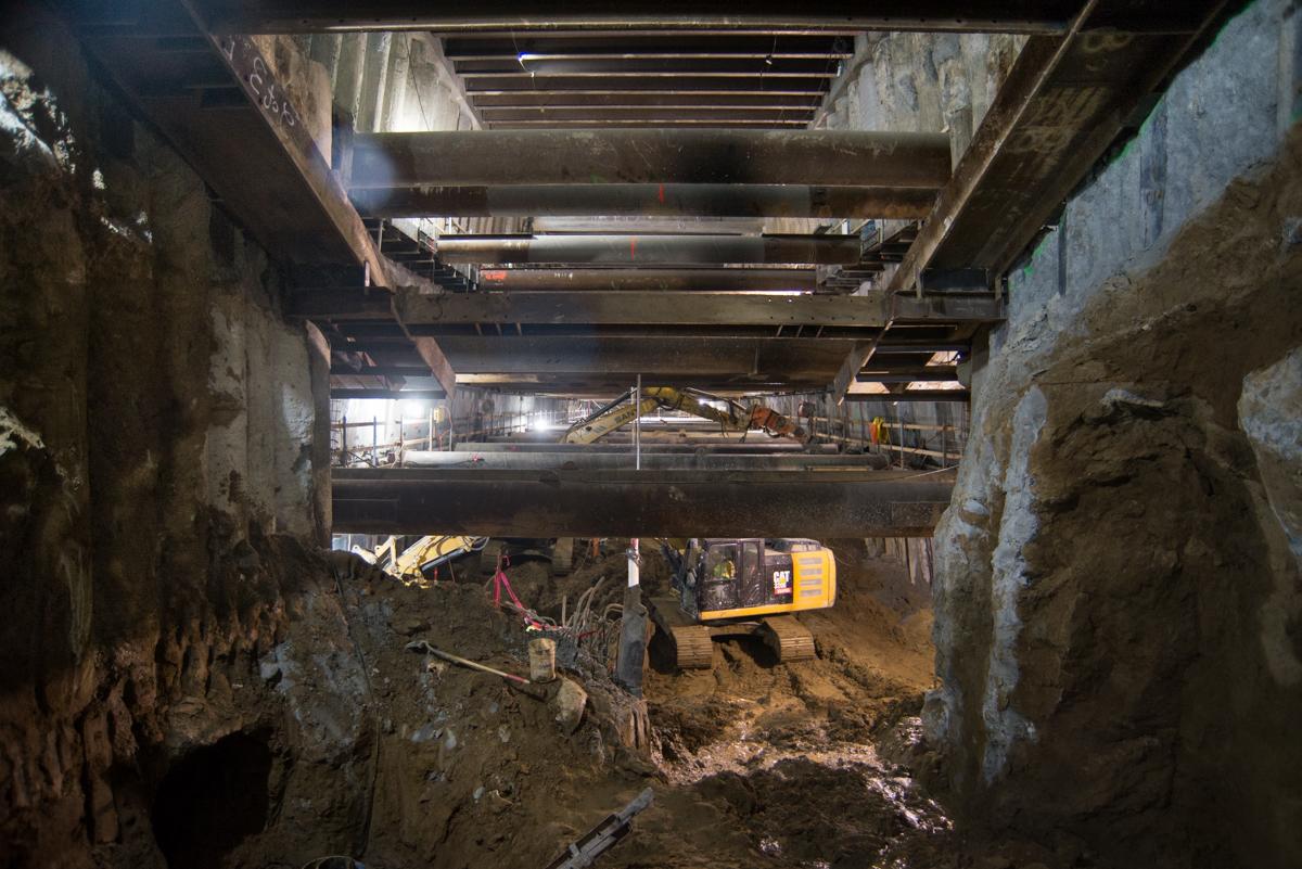 Construction site showing underground tunnel with concrete walls, steel beams, and yellow excavation equipment during Central Subway project.