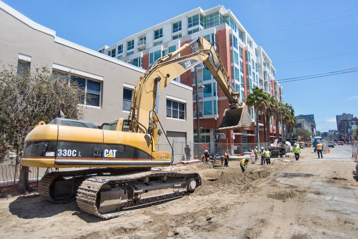 CAT excavator at 4th and Welsh worksite during Central Subway construction, with workers and modern buildings visible.