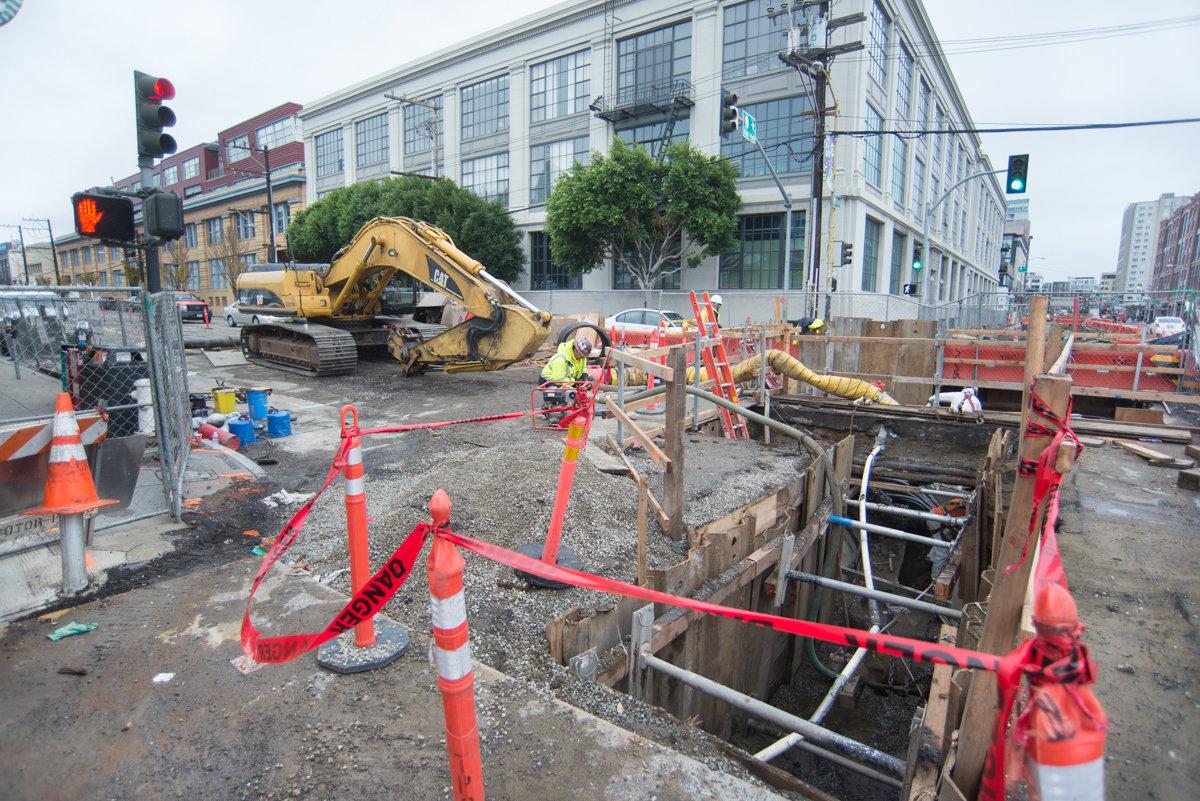 Construction site with excavator, safety barriers, and utility work at urban intersection near buildings.