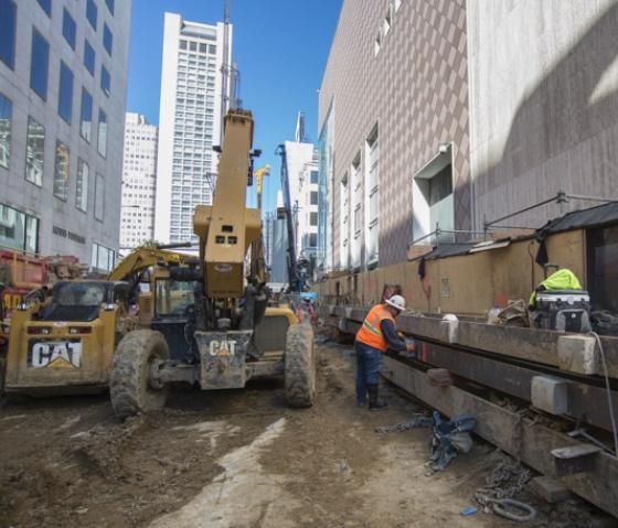 Construction equipment and workers prepare steel I-beams on urban street surrounded by tall buildings.