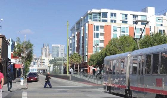 Light rail train at street-level station with modern residential buildings and city skyline in background.