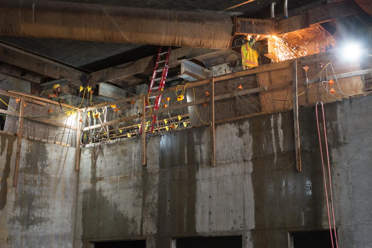 Workers on elevated platform performing nighttime construction inside concrete structure with steel bracing and equipment.