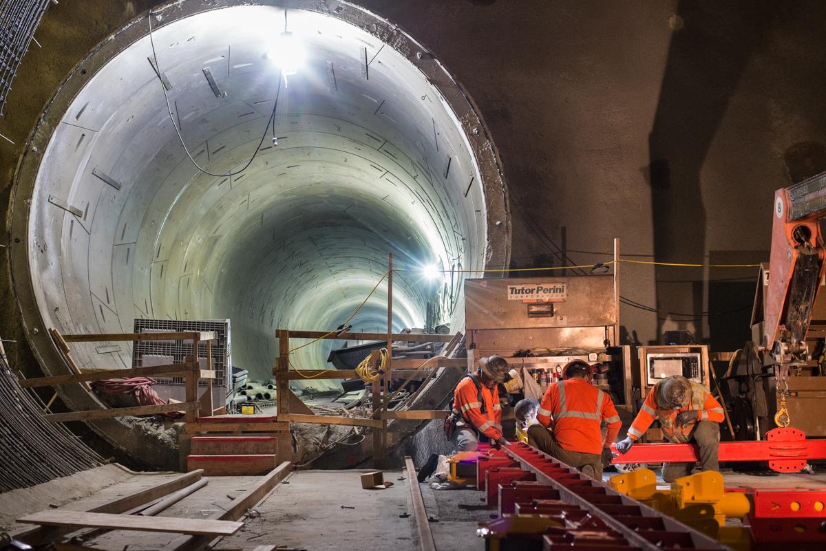 Workers in safety vests assemble track equipment inside a large underground tunnel cavern during transit construction.