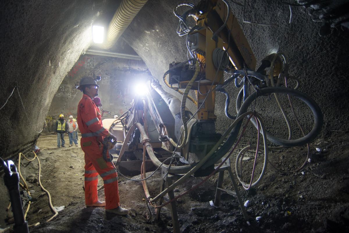 Worker in red suit operating shotcrete equipment inside underground transit tunnel excavation with bicycles and crew visible.