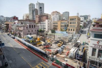 Aerial view of Chinatown construction site with excavation pit, red transit vehicle, surrounding buildings, and downtown skyline.
