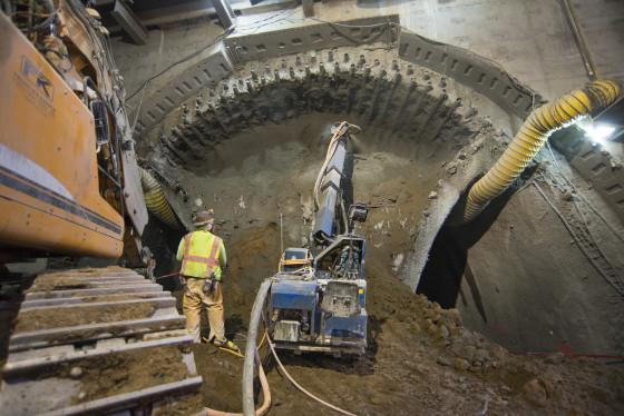 Worker in yellow safety vest inside tunnel excavation with large circular tunnel boring machine and construction equipment.