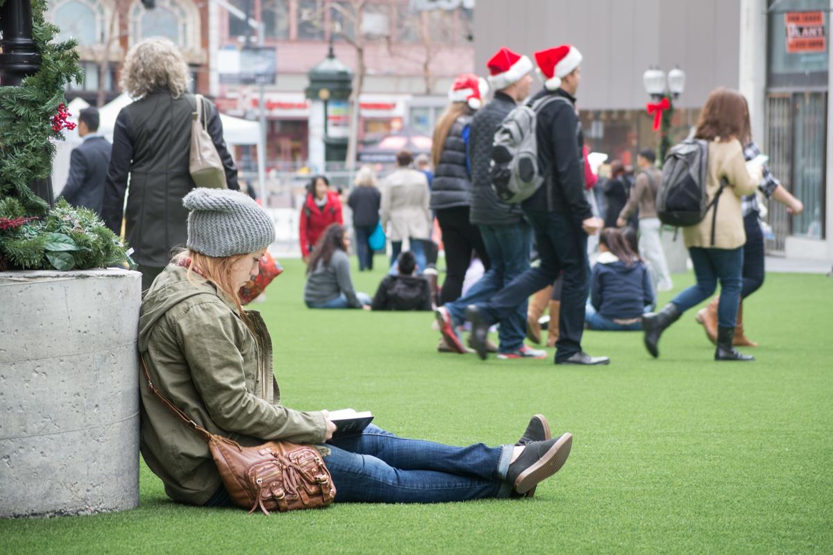 Woman sitting alone on green turf reading while holiday shoppers and pedestrians walk nearby in urban plaza.