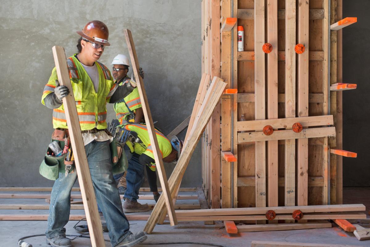Two construction workers in safety gear assemble wooden framing and forms inside a transit station.