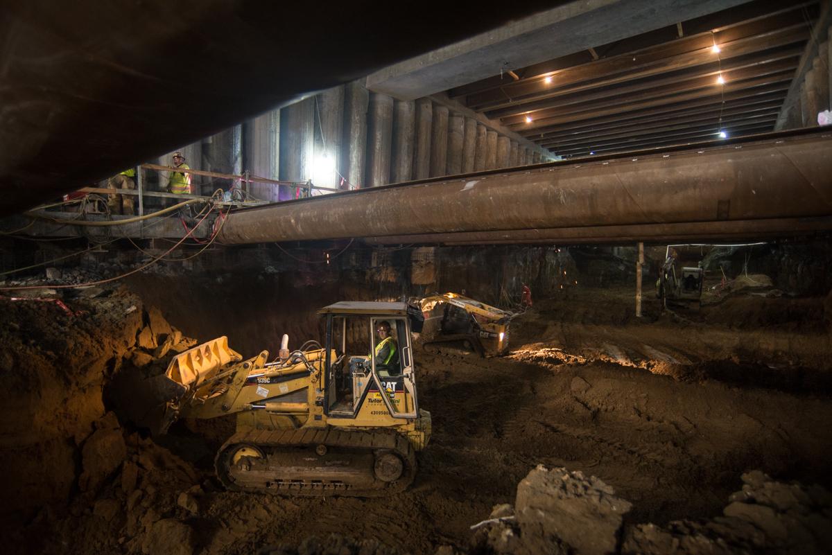Excavator working at night under a large pipe during Central Subway station construction.