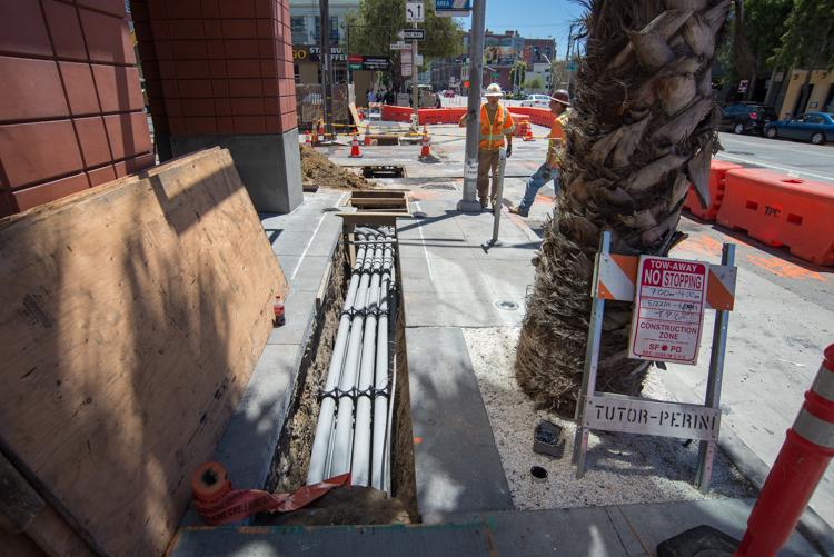 Construction site showing utility conduit installation in trench along sidewalk with palm tree and red barriers.