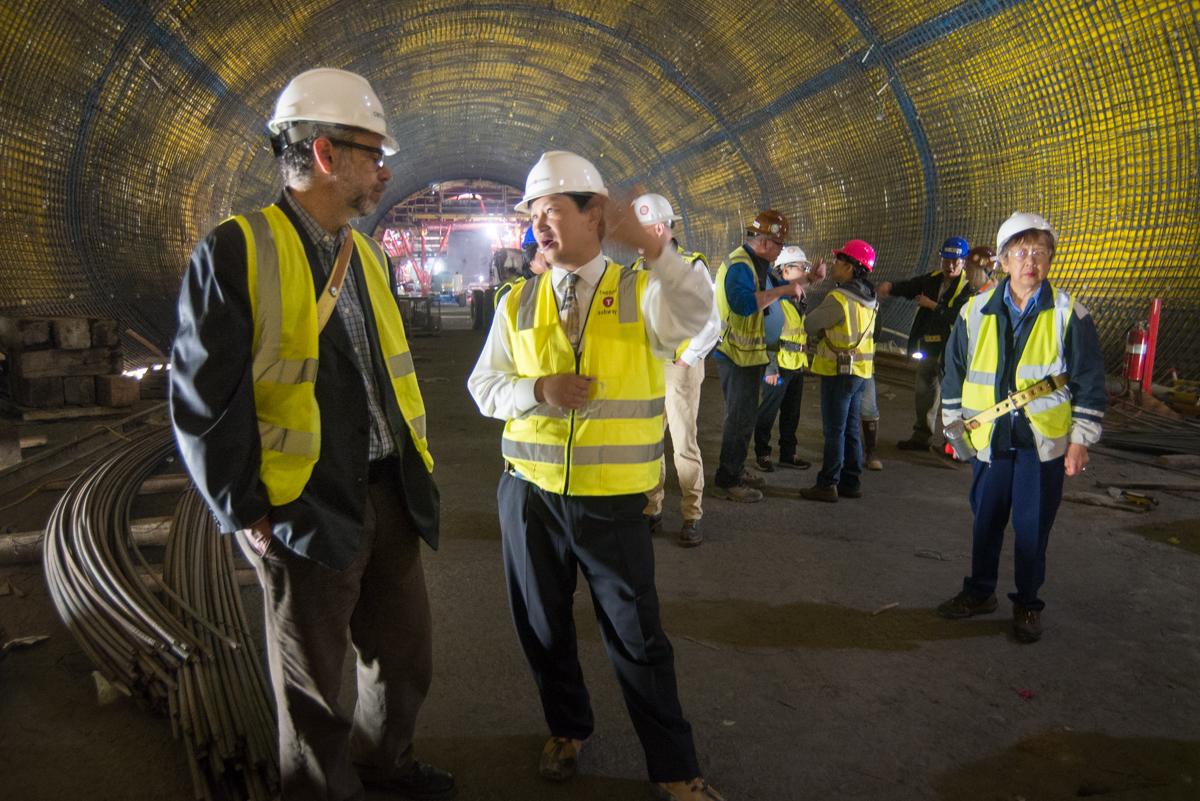 Workers and officials in hard hats and safety vests tour a large underground tunnel with curved yellow walls and railroad tracks.