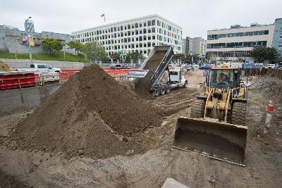 Construction site with excavator and dump truck moving soil pile near urban buildings.