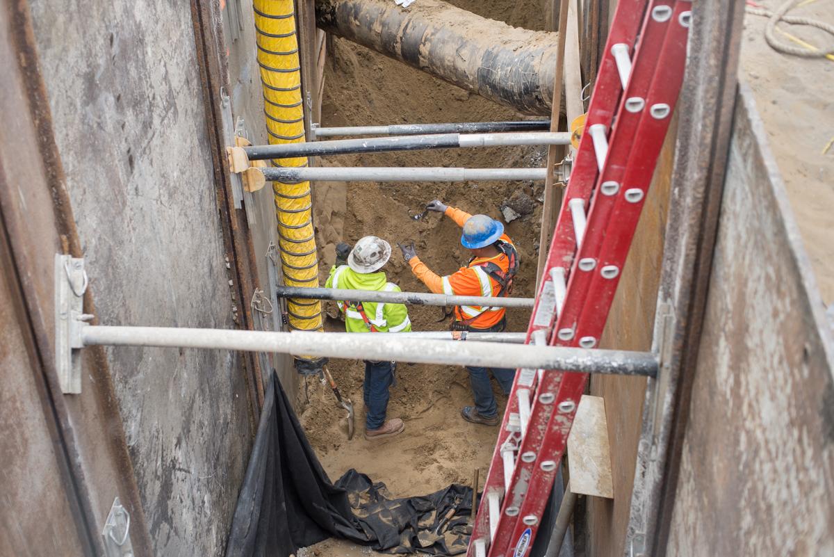 Workers in safety gear install sewer line in deep trench near Yerba Buena Moscone Station.