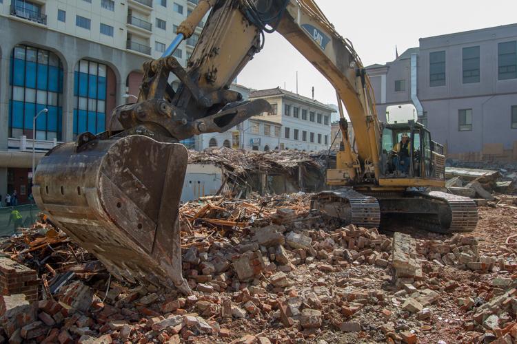 Excavator with bucket demolishing brick building at urban construction site surrounded by debris