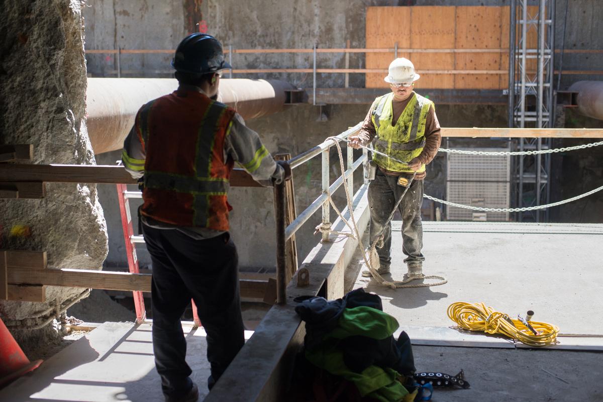 Two construction workers in safety vests coordinate equipment lifting on a mezzanine platform at a transit station construction site.