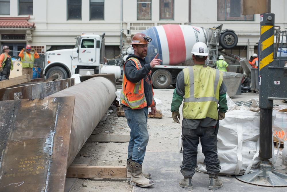 Construction workers in safety vests discuss operations at a transit station site with concrete pump truck and equipment visible.