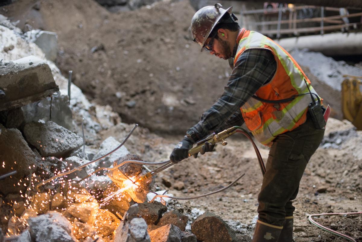Worker in safety gear using a cutting torch on rebar at underground transit construction site.