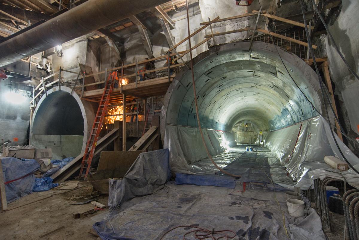 Central Subway tunnel under construction with steel reinforcement and concrete lining visible inside cylindrical tunnel section.