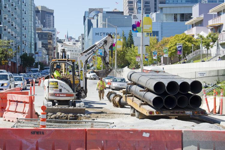 Construction site on 4th Street with excavator, large pipes, and traffic controls in urban setting.