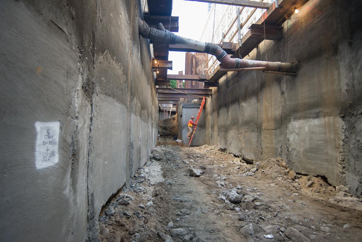 Narrow underground passage with concrete walls, steel pipes overhead, and construction debris on ground floor.