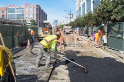 Workers in safety vests rake and smooth asphalt on an urban street construction site surrounded by green fencing.