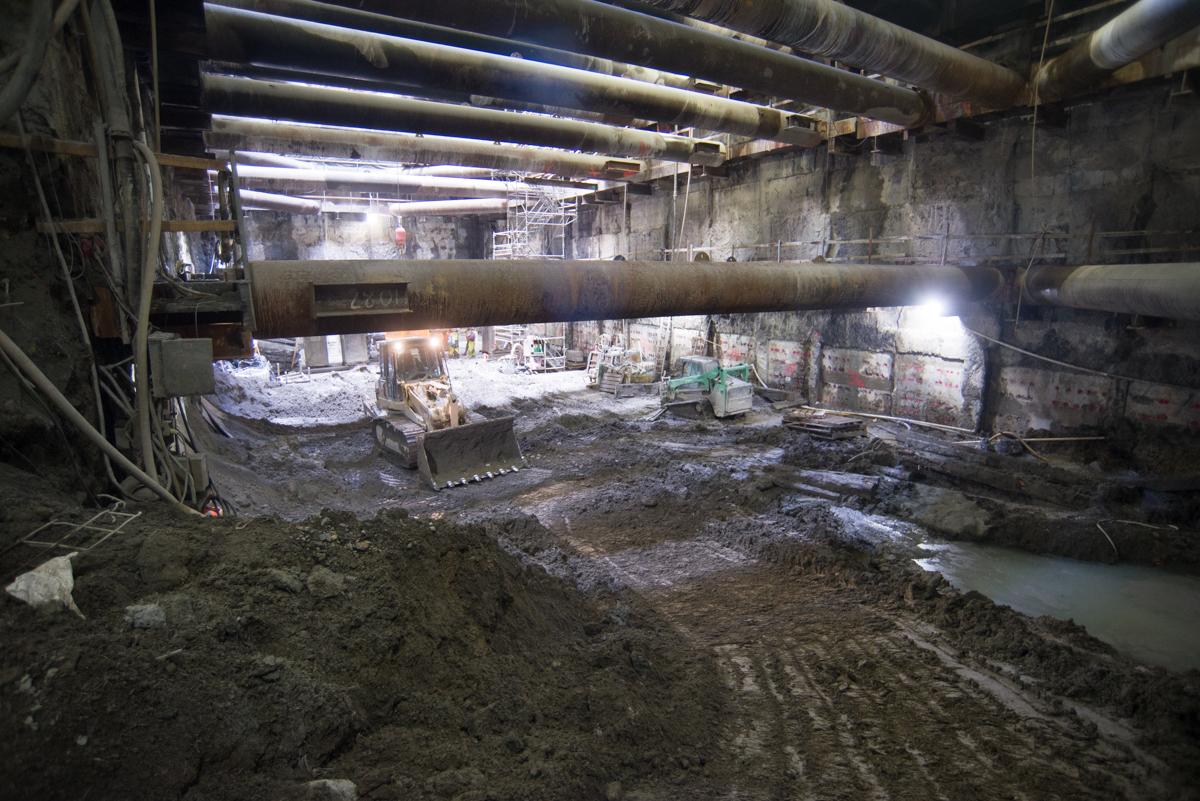 Underground transit tunnel under construction with excavator, bulldozer, and exposed concrete ceiling beams.