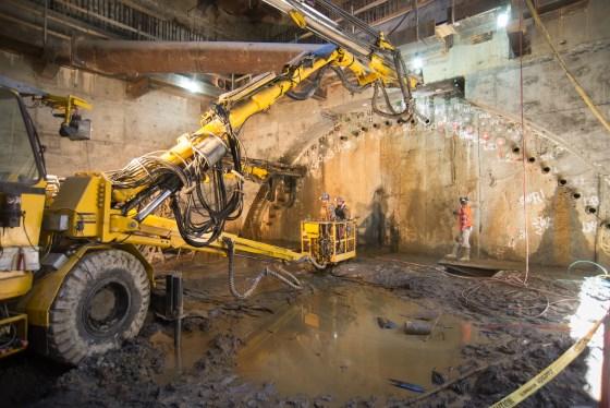 Yellow drilling rig operates inside underground construction site with workers and wet ground visible.