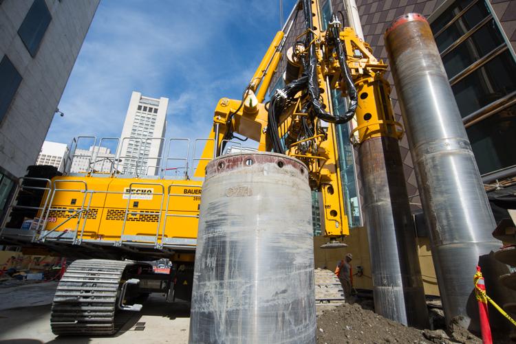 Yellow drilling rig installing pilings on urban street with concrete cylinders and city buildings in background.