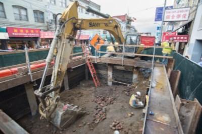 Excavator removing soil from underground construction pit on city street with buildings and Chinese signage visible.