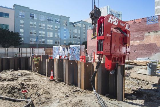 Red vibro hammer machine driving steel sheet piles into ground at construction site with apartment building in background.