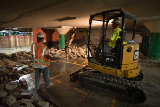 Worker in safety vest operates small excavator during underground construction and demolition work.
