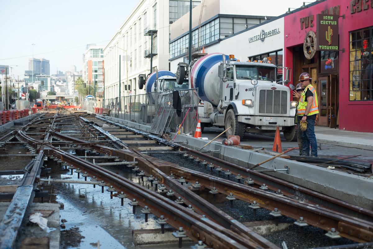 Concrete truck with mixer drum positioned over railroad tracks during foundation work at 4th and Brannan Station construction site.