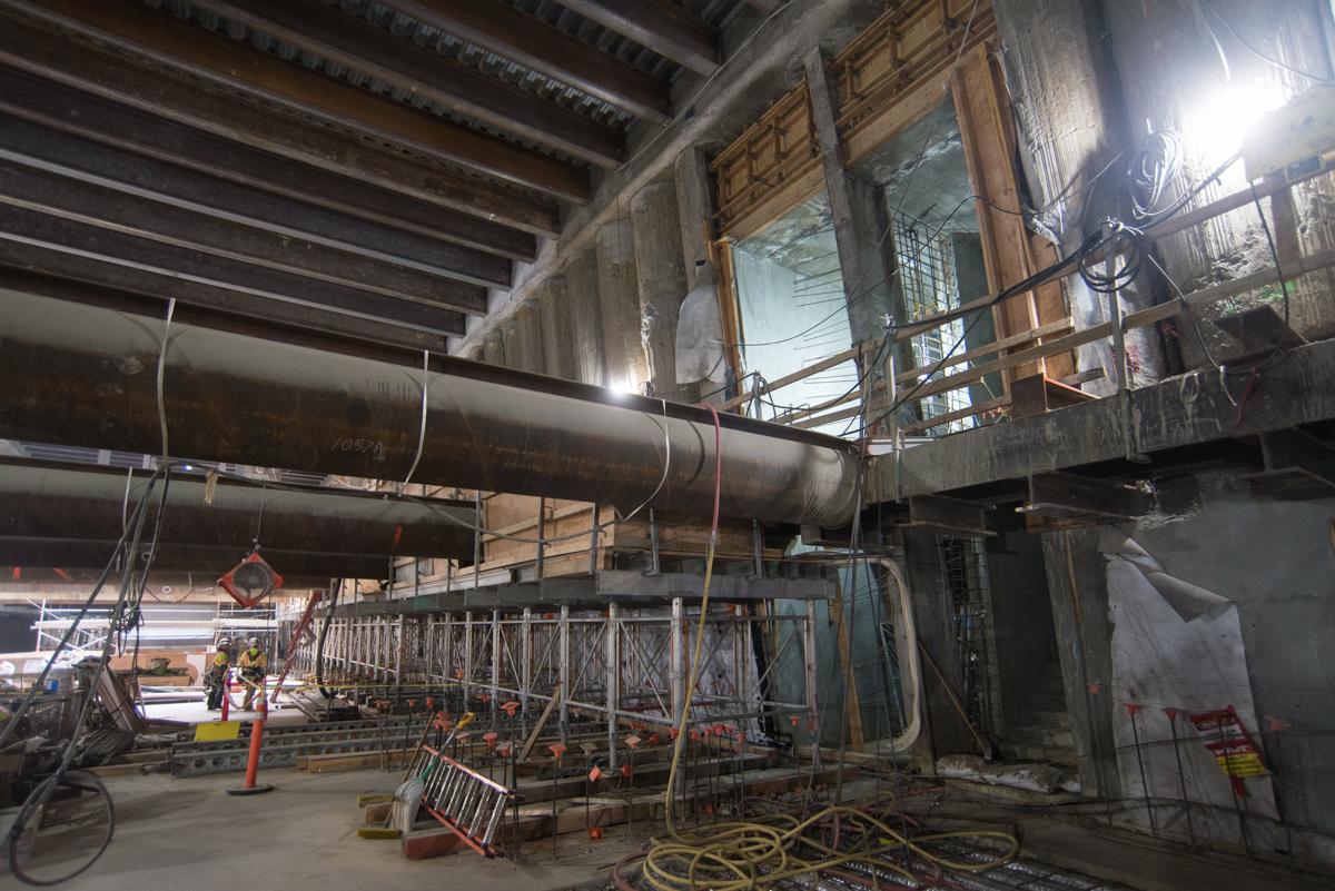 Construction work inside station box with large cylindrical pipe, metal scaffolding, and structural formwork under concrete ceiling.