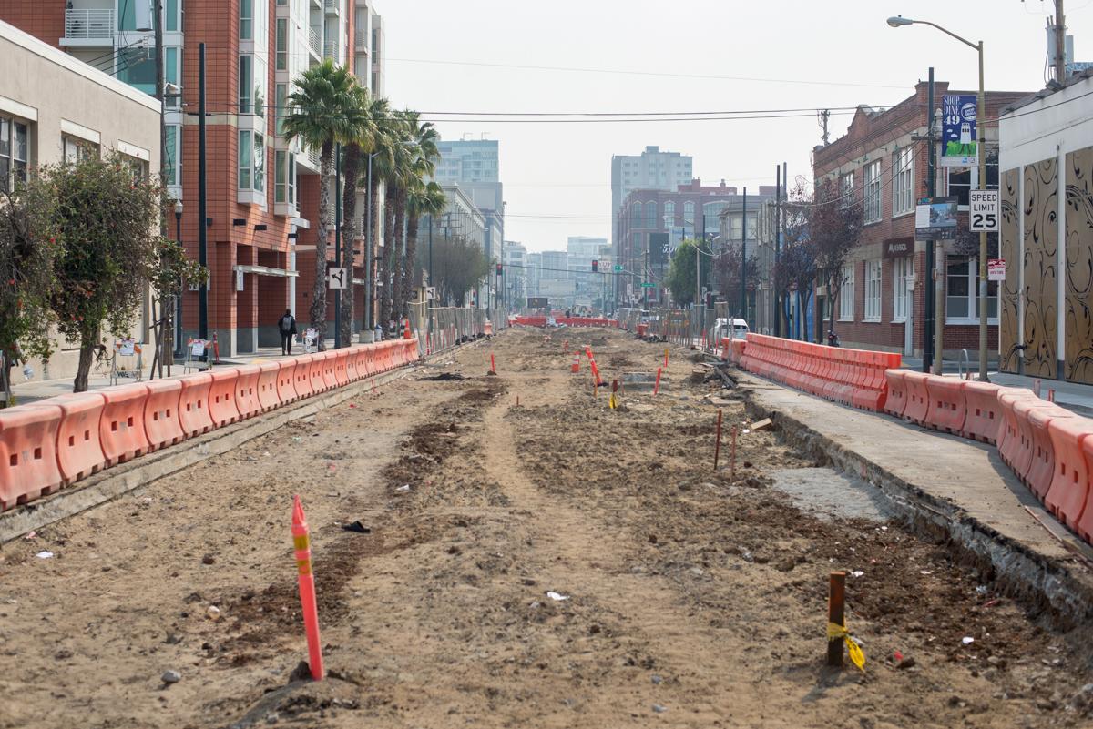 Construction site on 4th Street with exposed earth, orange barriers, and downtown buildings in background.