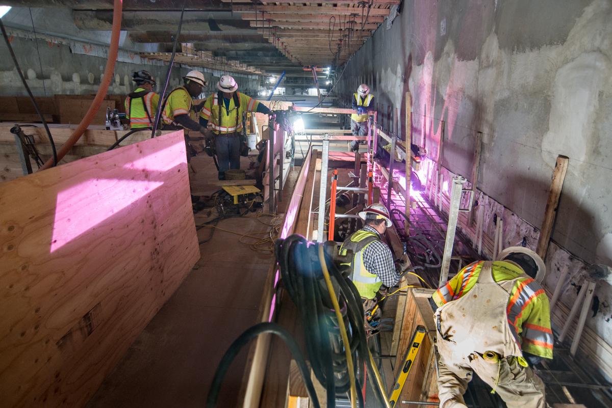 Workers in safety gear inspect construction materials and equipment in an underground transit tunnel with pink lighting.