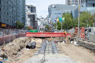 Construction site with excavated trench, sewer pipes, and red equipment along urban street.