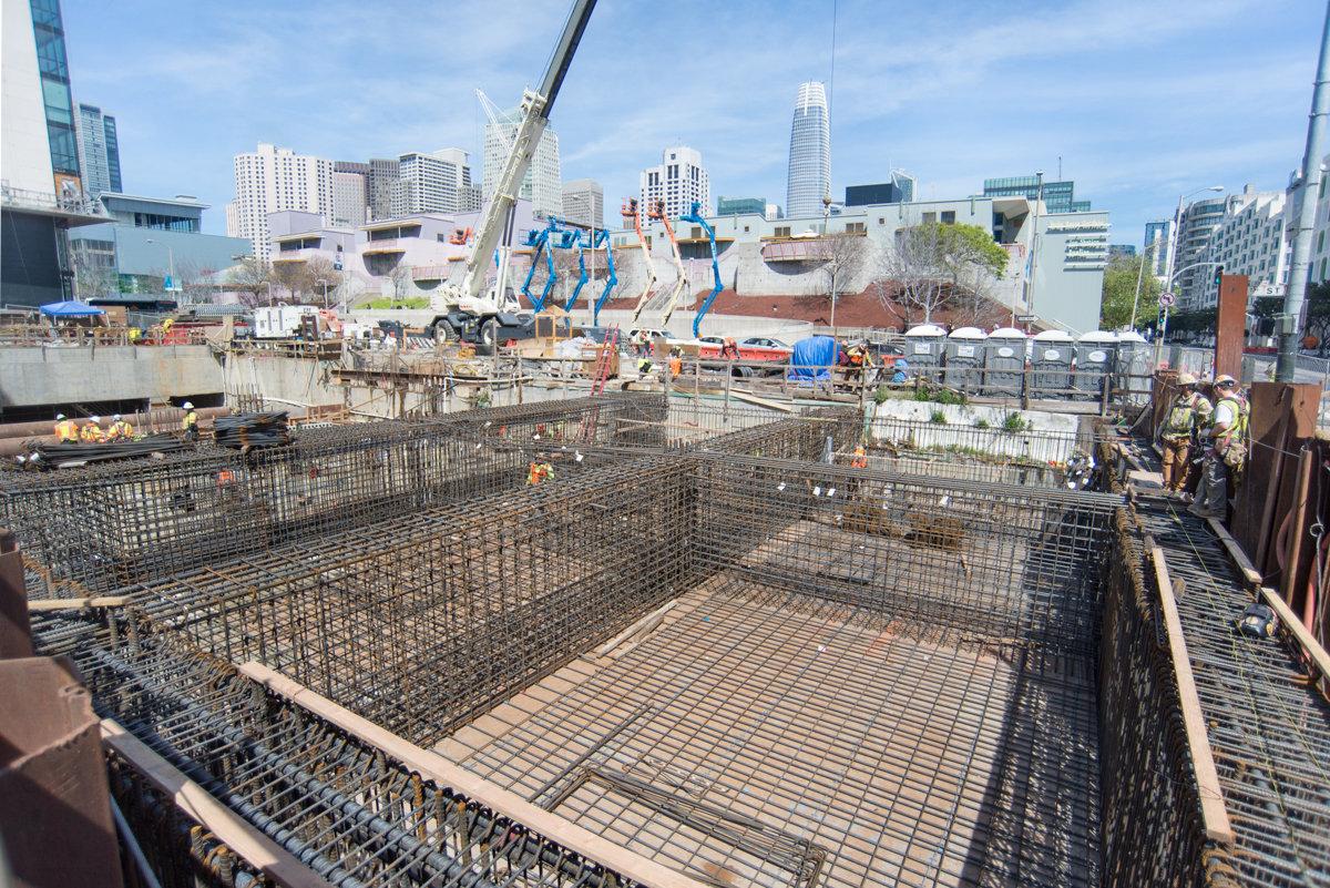 Construction site with reinforced steel rebar cages and concrete framework for Central Subway project, San Francisco skyline in background.