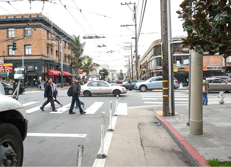 Pedestrians cross at intersection while white posts and beige zone protect curb area from vehicle traffic.