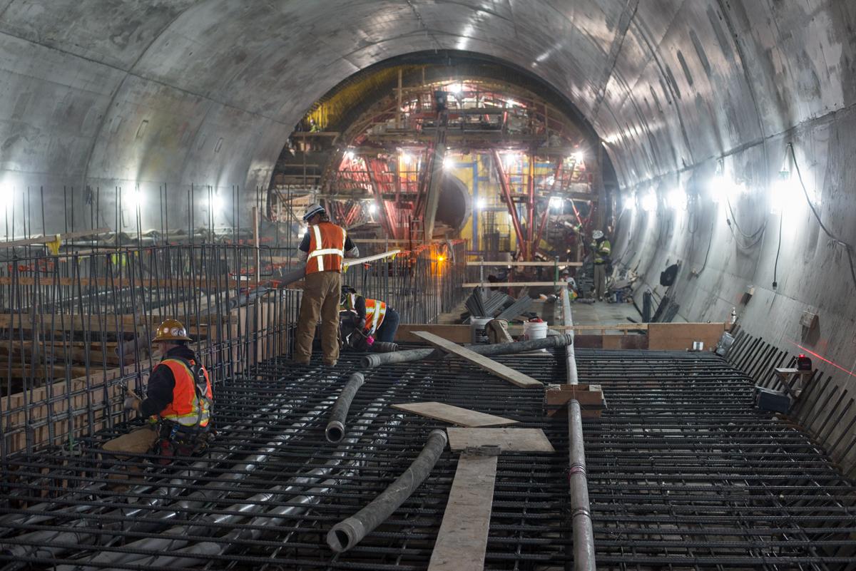 Workers install rebar and conduits inside a tunnel under construction for a transit platform.