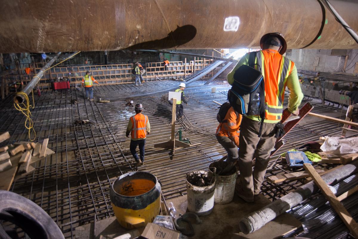 Construction workers on reinforced concrete deck inside station box with rebar and equipment visible.