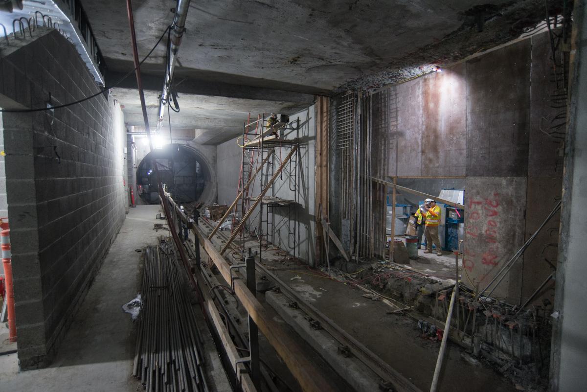 Underground transit station under construction with workers, scaffolding, and exposed infrastructure along platform.