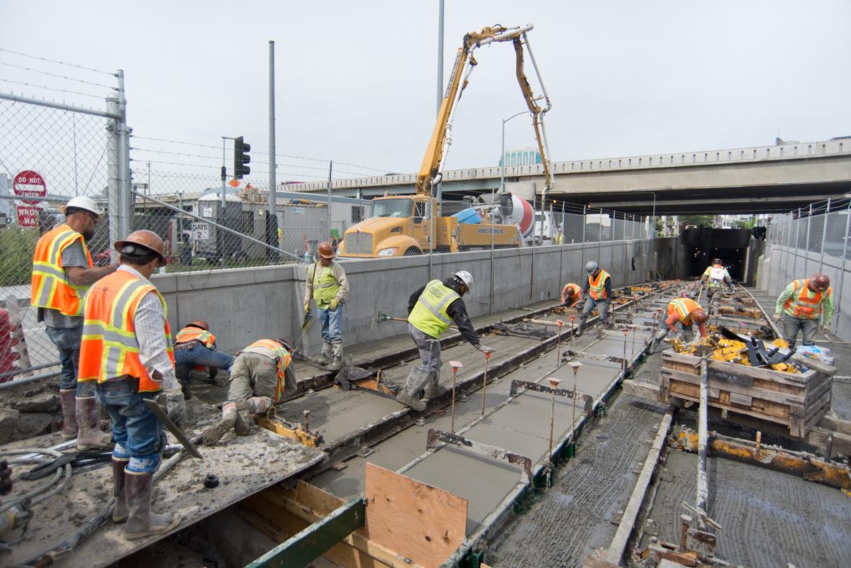 Construction workers pour concrete for tunnel portal foundation at Central Subway project site under highway overpass.