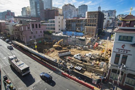 Construction site with excavation equipment and workers at Stockton Street in urban setting with buildings and street traffic.